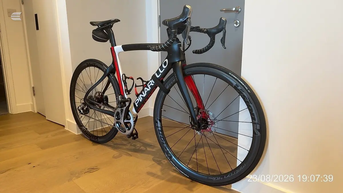 Black, blue and red Pinarello road bike on wooden floor indoors, drive side view, against white walls and grey door.