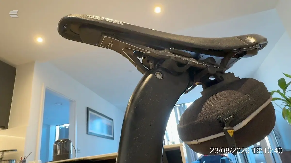 Black stationary exercise bike indoors, non-drive side, against blue wall with recessed lighting and potted plant visible.