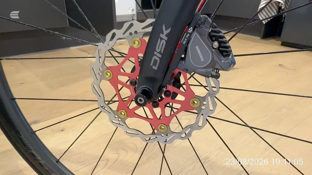 Close-up of disc brake system on red mountain bike, front wheel with metallic rotor and caliper, wooden floor background, drive side angle.