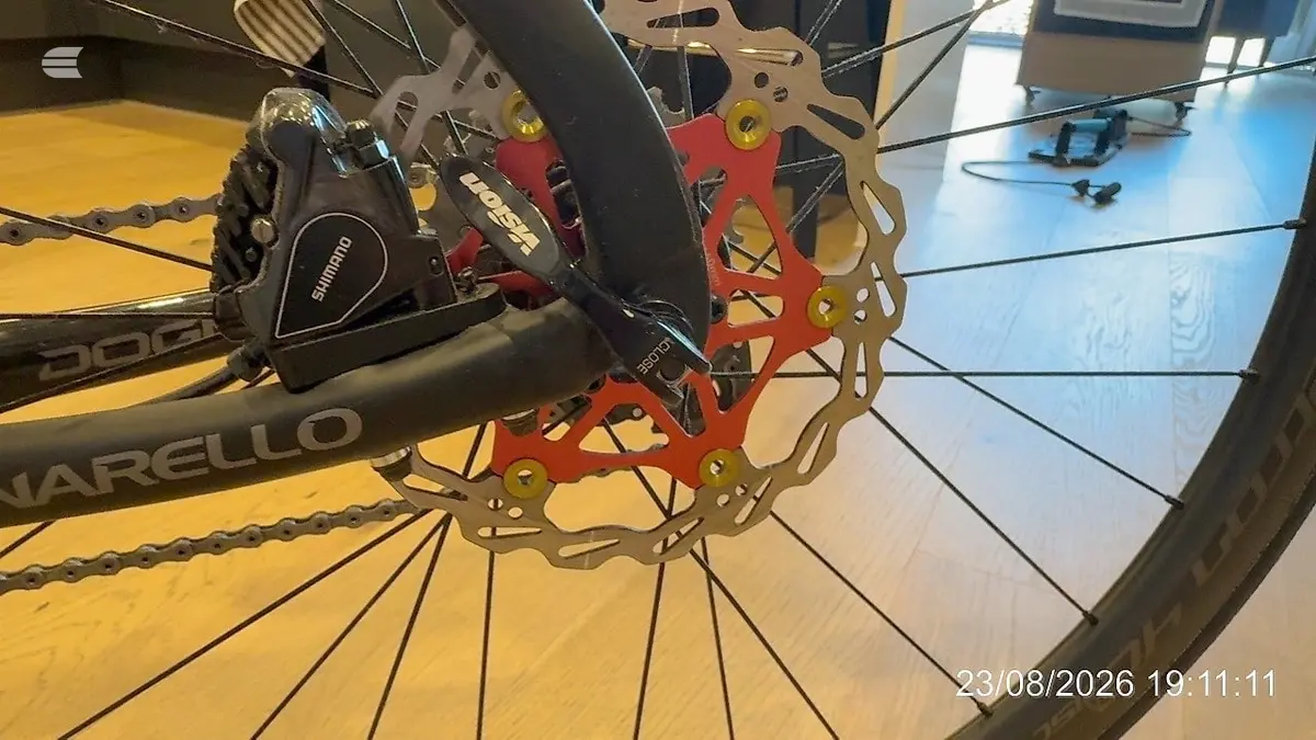 Close-up of disc brake system on a Marello mountain bike, showing red rotor and Shimano caliper, drive side, indoors on wooden floor.