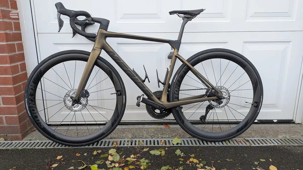 Gold and black gravel or endurance road bike on driveway, drive side, against white garage door and red brick wall.