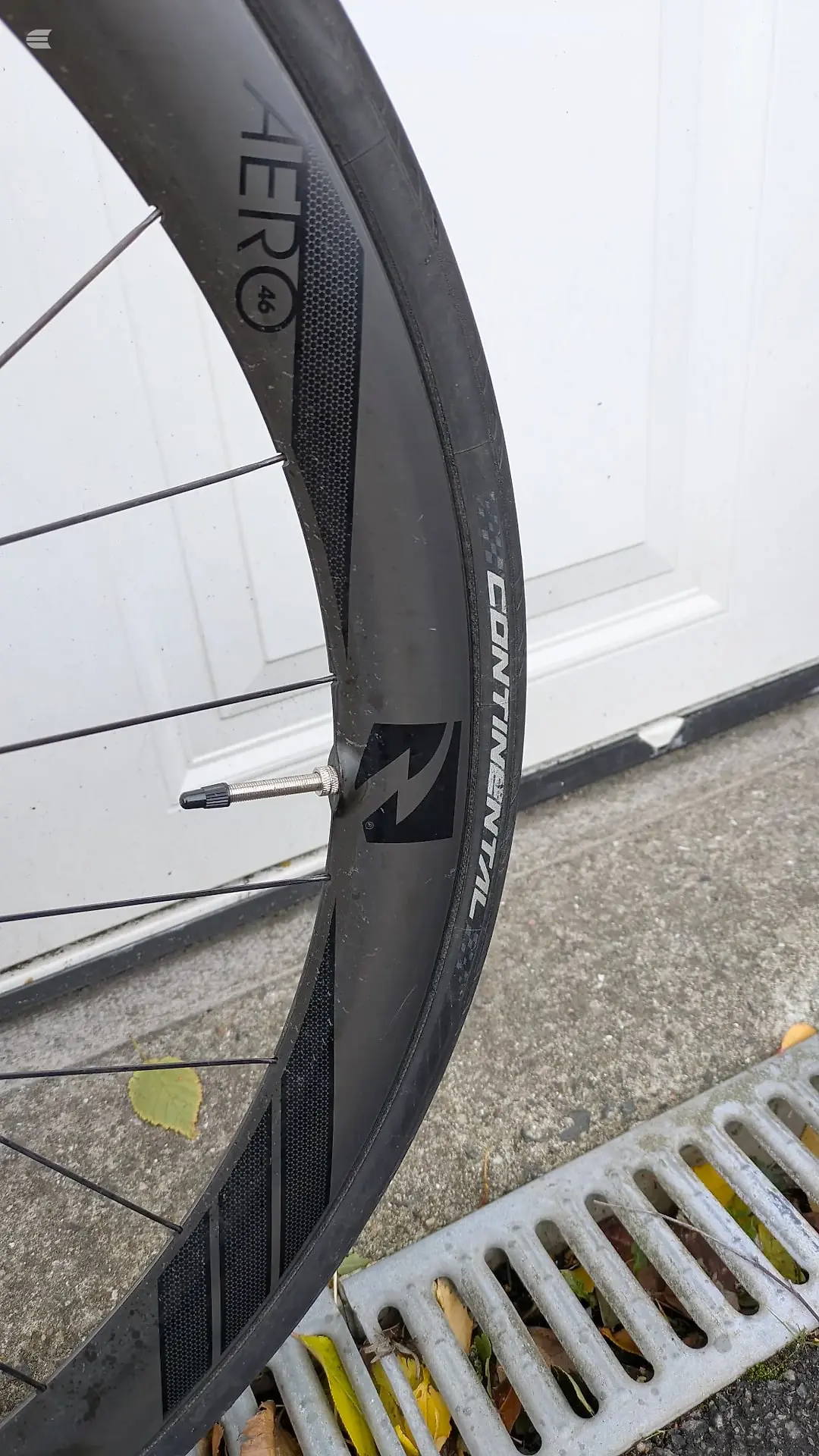 Close-up of black carbon fibre road bike wheel with Zipp Firecrest rim branding, drive side, against white garage door and grey pavement.