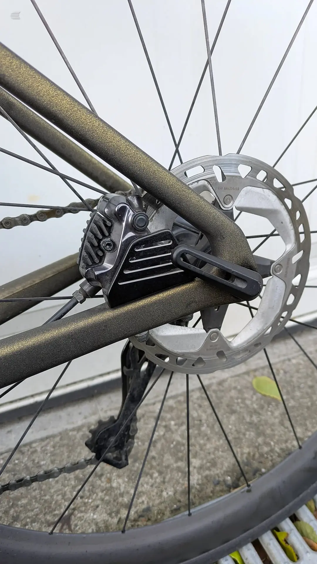 Close-up of disc brake caliper and rotor on olive-green gravel bike, drive side, against pavement and sky.