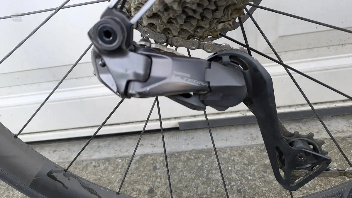 Shimano rear derailleur and chain on a bicycle, drive side close-up, against concrete ground and white metal shutter.