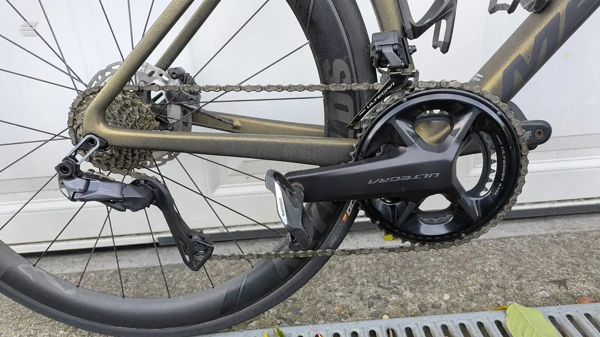Gravel bike drivetrain detail, drive side, showing cassette, chain, derailleur and crankset on pavement with metal shutter background.