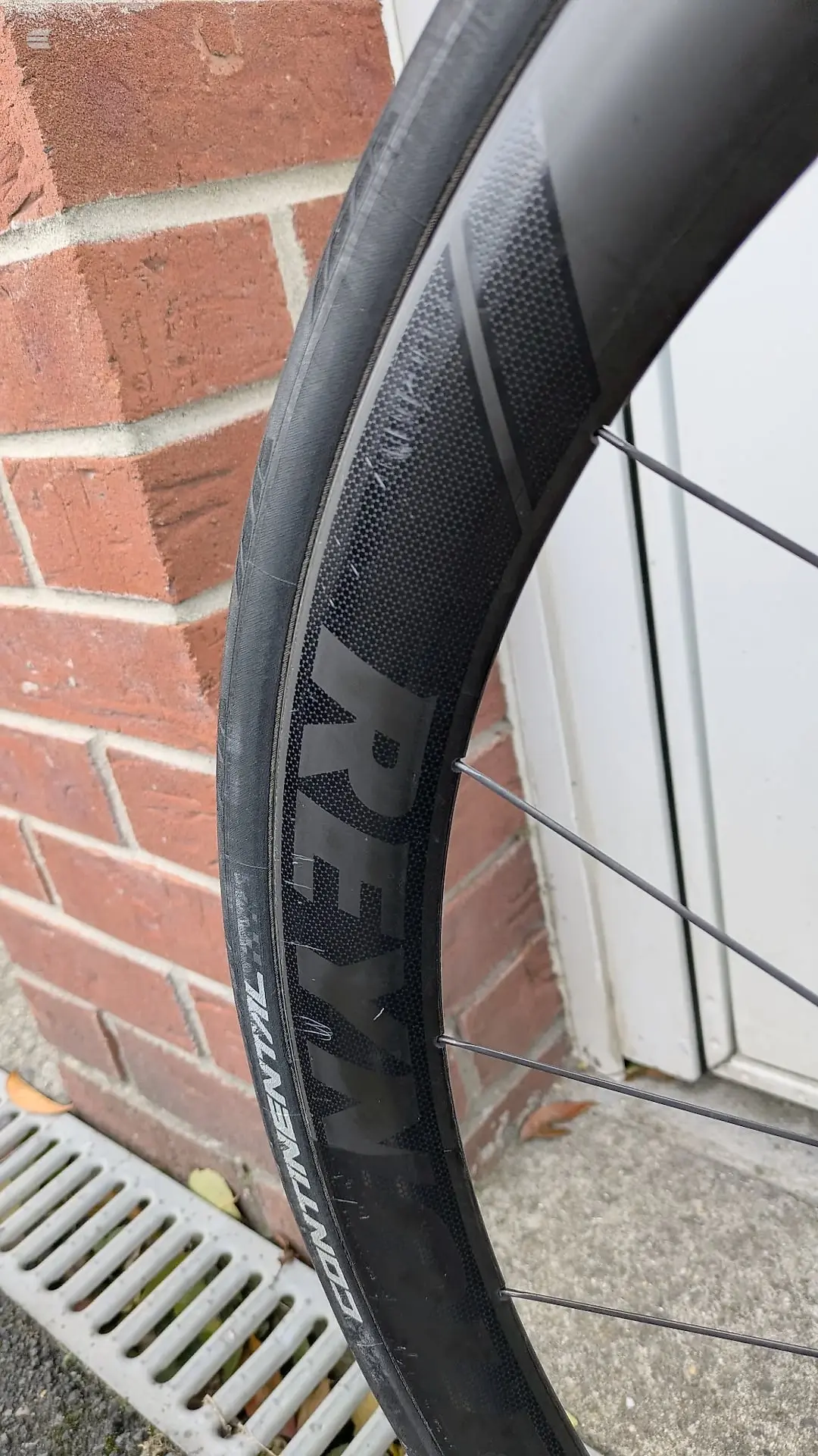 Close-up of a black carbon fibre bicycle wheel rim with visible brake surface and spoke detail, leaning against a red brick wall with white trim.