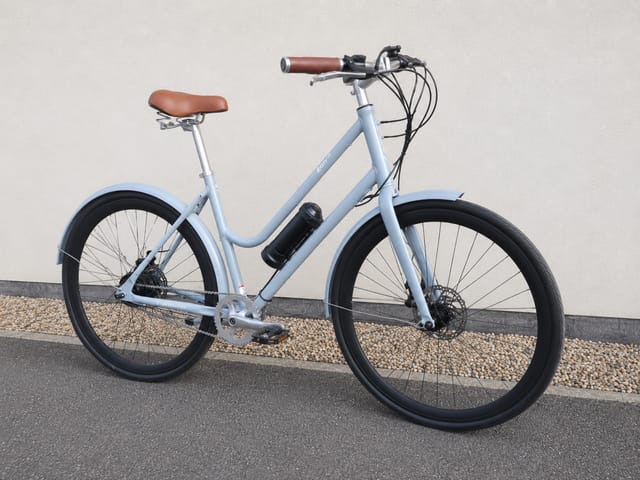 Light blue step-through e-bike with brown leather saddle and grips, disc brakes, and black fenders, parked on gravel against a white wall, drive side view.