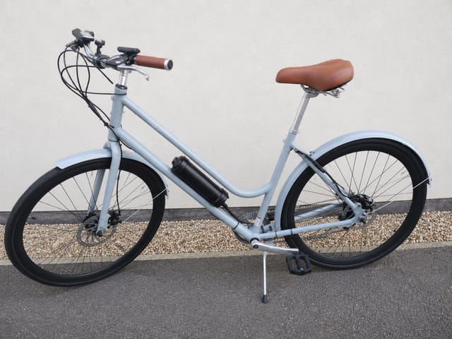 Light blue step-through e-bike with brown saddle and grips, black wheels with mudguards, kickstand deployed, parked on asphalt against gravel and white wall,
