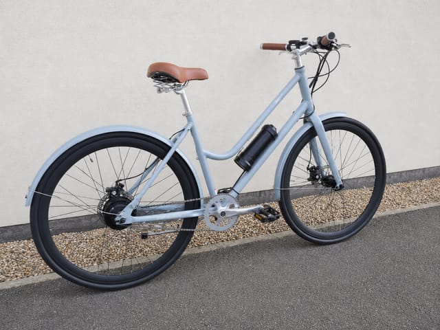 Light blue step-through e-bike with brown saddle and grips, black mudguards and tyres, parked on gravel against a white wall, drive side view.