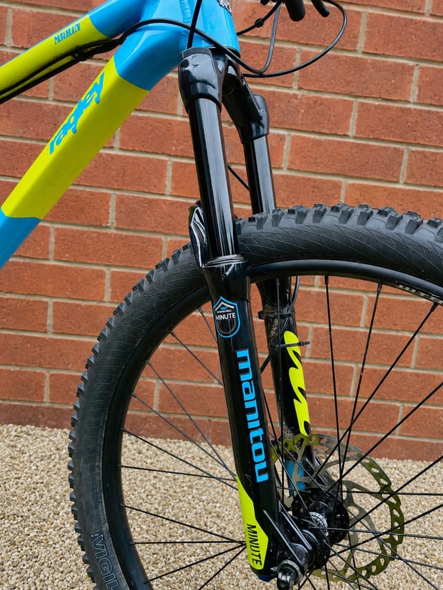 Blue and yellow hardtail mountain bike, front wheel close-up against red brick wall, drive side, showing black tyre and suspension fork.