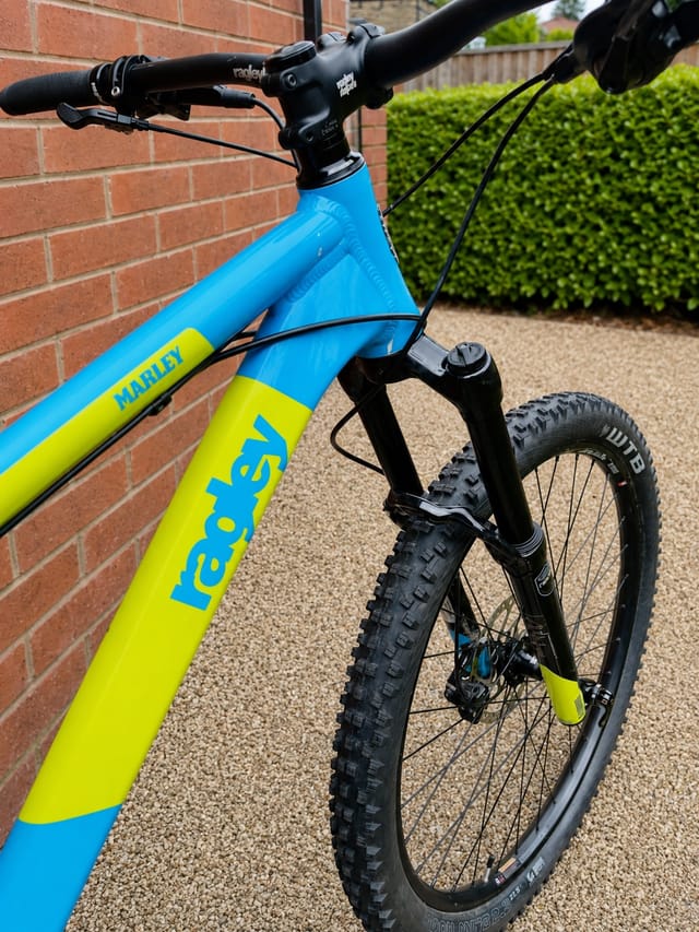 Blue and yellow Nandi hardtail mountain bike, drive side, leaning against red brick wall with gravel ground and hedge background.