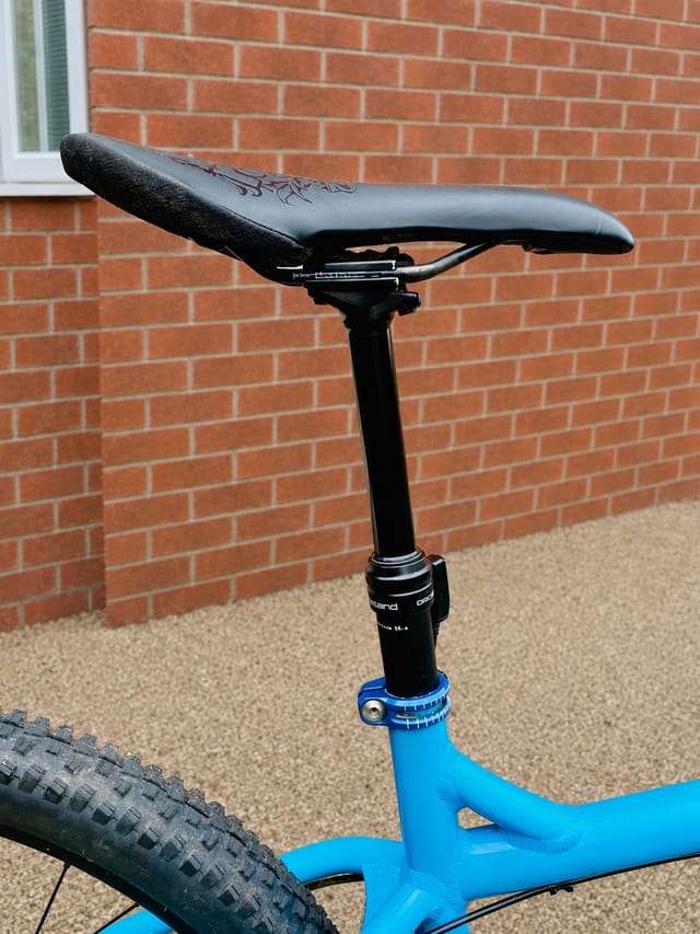 Black seatpost with blue clamp and black saddle on bright blue mountain bike frame against red brick wall.