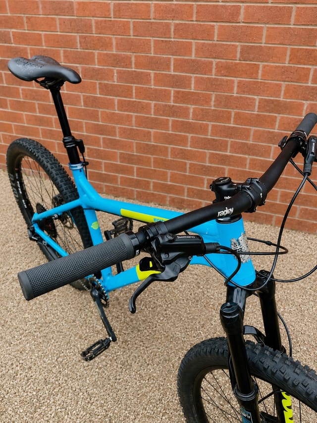 Blue and yellow hardtail mountain bike with flat handlebars, three-quarter view from drive side, against a red brick wall.