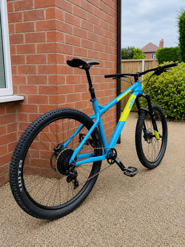 Blue and yellow hardtail mountain bike on concrete driveway, drive side, against red brick house wall.