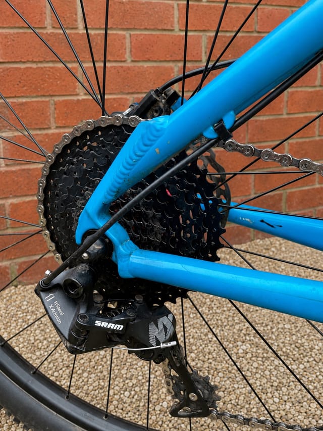 Close-up of blue mountain bike rear drivetrain, drive side, showing SRAM derailleur, cassette, and chain against brick wall background.