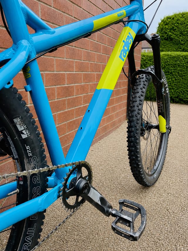 Blue and yellow hardtail mountain bike against a brick wall, three-quarter drive-side view. Front fork and headtube in focus, rear wheel visible.