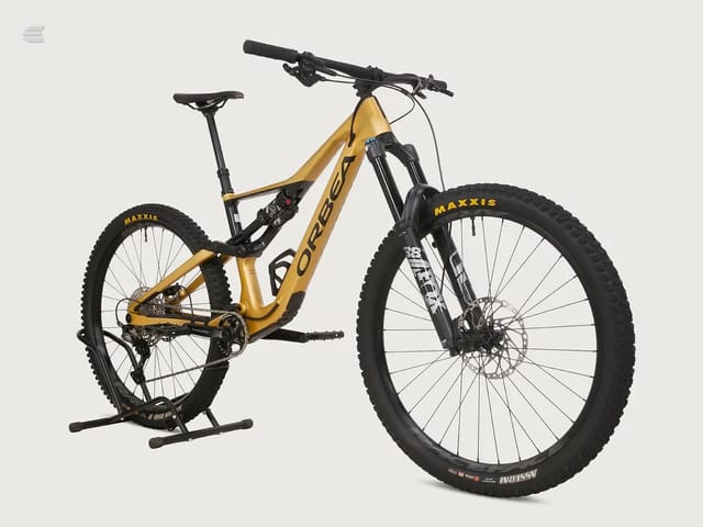 Gold and black Orbea full-suspension mountain bike on a stand, drive side, against a plain grey background.