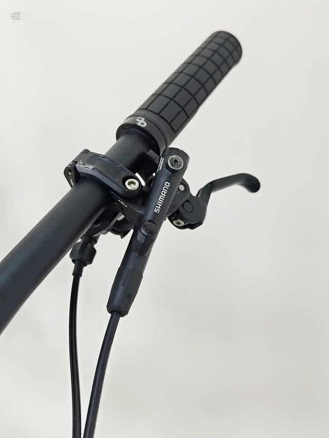 Shimano hydraulic disc brake lever and grip detail on black handlebar, close-up against plain background.