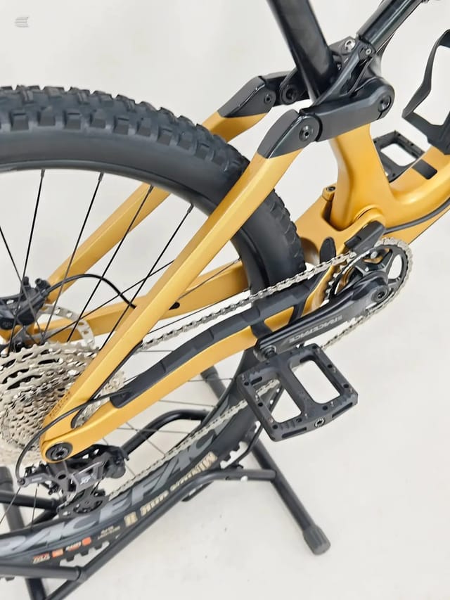 Yellow gravel bike on a stand, drive side, showing drivetrain, chain, cassette, rear derailleur, pedals and lower frame detail against a white background.