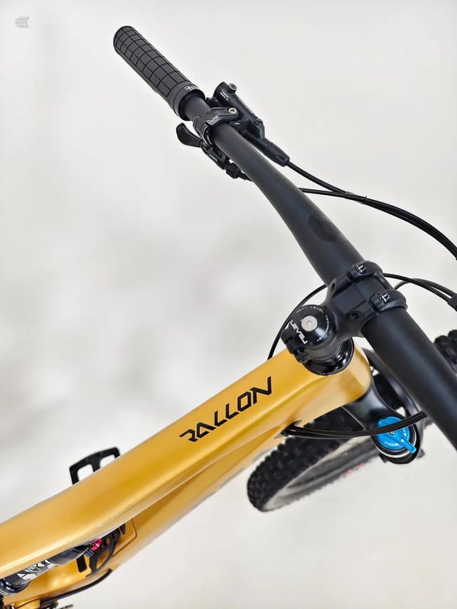 Gold Rallon scooter or kick bike with black handlebars and blue front wheel, angled upward against white sky, non-drive side view.