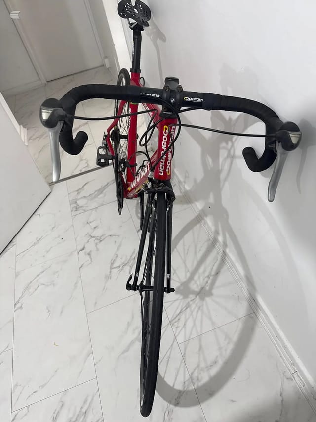 Red and black road bike with drop bars, viewed from above against a marble floor and white wall, drive side visible.