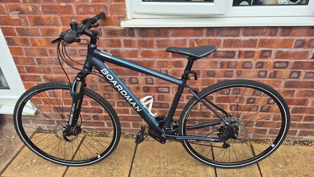 Black and blue Boardman hybrid bike on concrete paving, non-drive side, against a red brick wall with white window frame above.