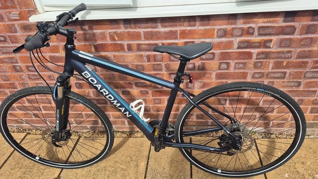 Boardman hybrid bike, black and blue frame, non-drive side, parked against red brick wall on paved ground.