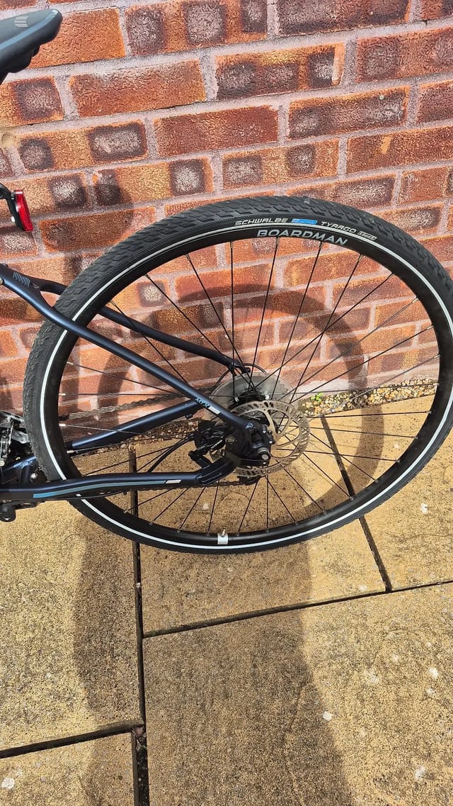 Schwalbe Tyranno 28 tyre on rear wheel of blue hybrid bike, drive side, leaning against red brick wall on paved ground.