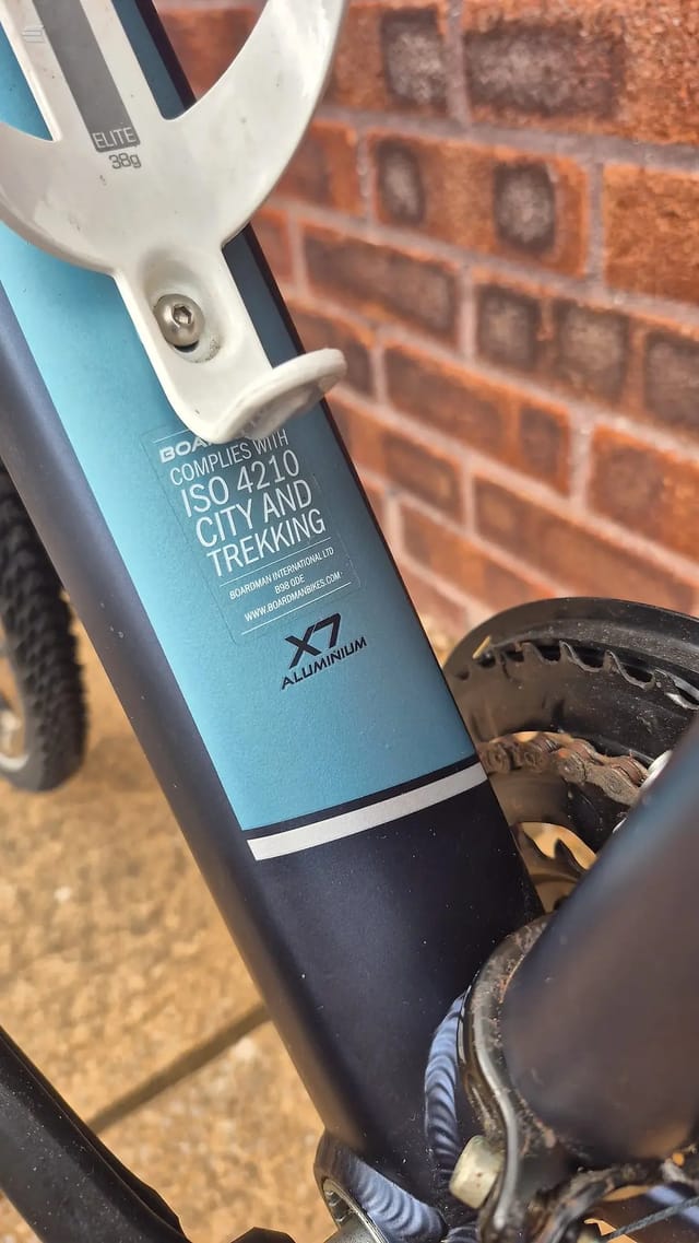 Close-up of blue and black aluminium city bike frame tube showing ISO 4210 compliance label and seat tube detail, against brick wall background.