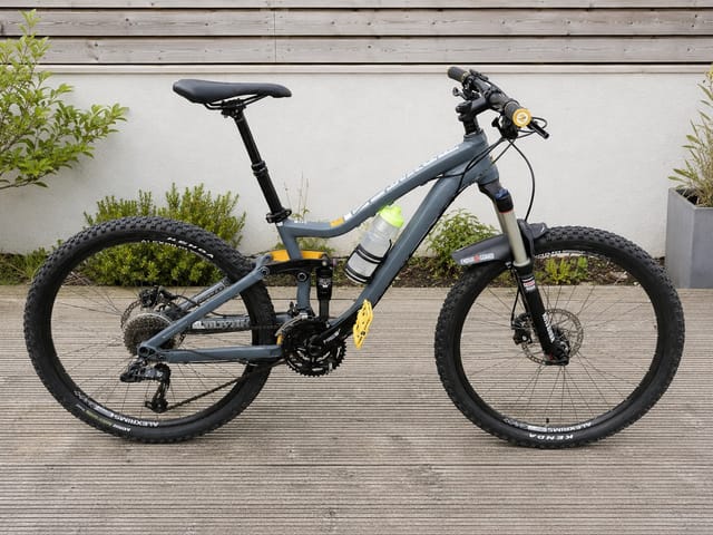 Grey and yellow full-suspension mountain bike on wooden decking, drive side, against white wall with potted plants.