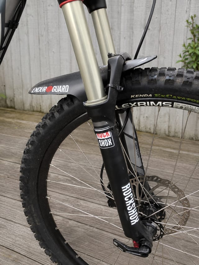 Black hardtail mountain bike with silver fork and red accents, front three-quarter view on wooden deck against concrete wall. Drive side visible.