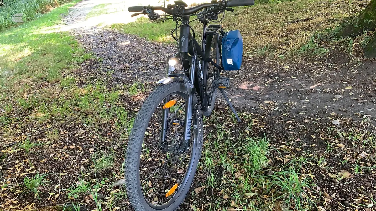 Black hardtail mountain bike on gravel path, drive side, with front light, blue pannier bag, and yellow reflectors visible.
