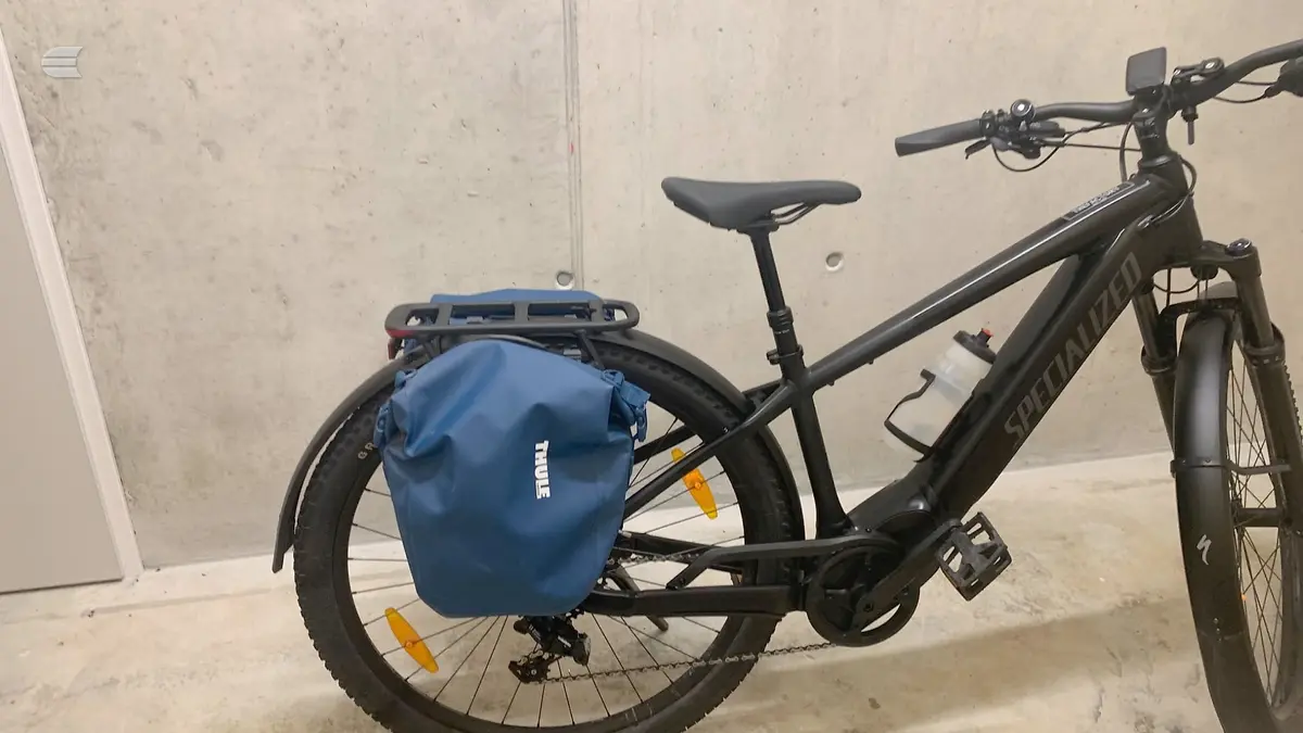Black step-through e-bike with blue Thule pannier bag on rear rack, drive side, parked against concrete wall indoors.