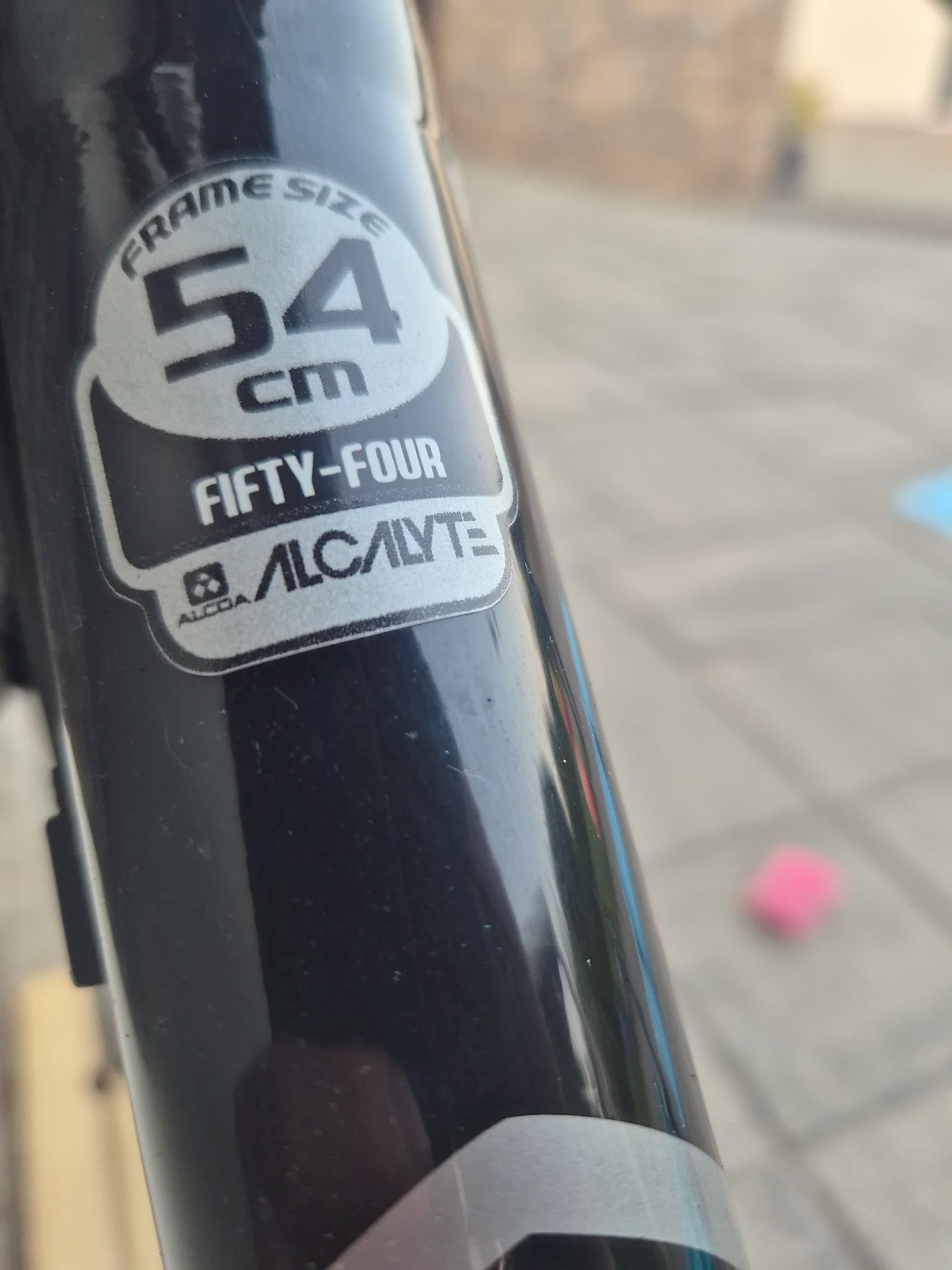 Close-up of black bicycle frame tube showing white decal label reading 'FRAMESIZE 54cm FIFTY-FOUR' and 'ALCAINTE' branding, outdoors on pavement.