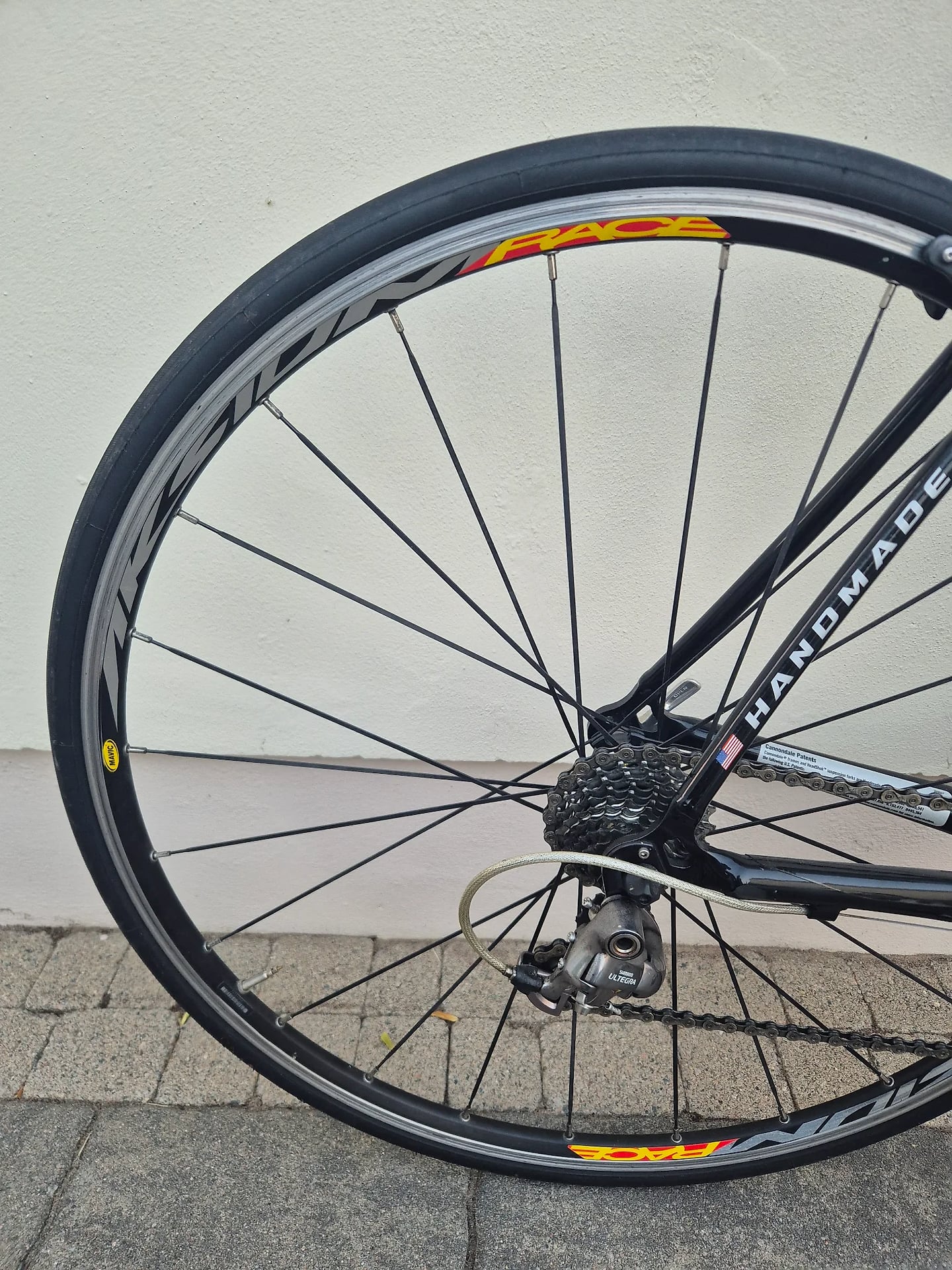 Rear wheel of a black Cannondale road bike with red and yellow rim decals, drive side, against a white wall and paved ground.