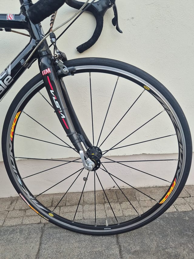 Black road bike front wheel with orange and yellow rim decals, drive side, leaning against white wall and grey pavement.