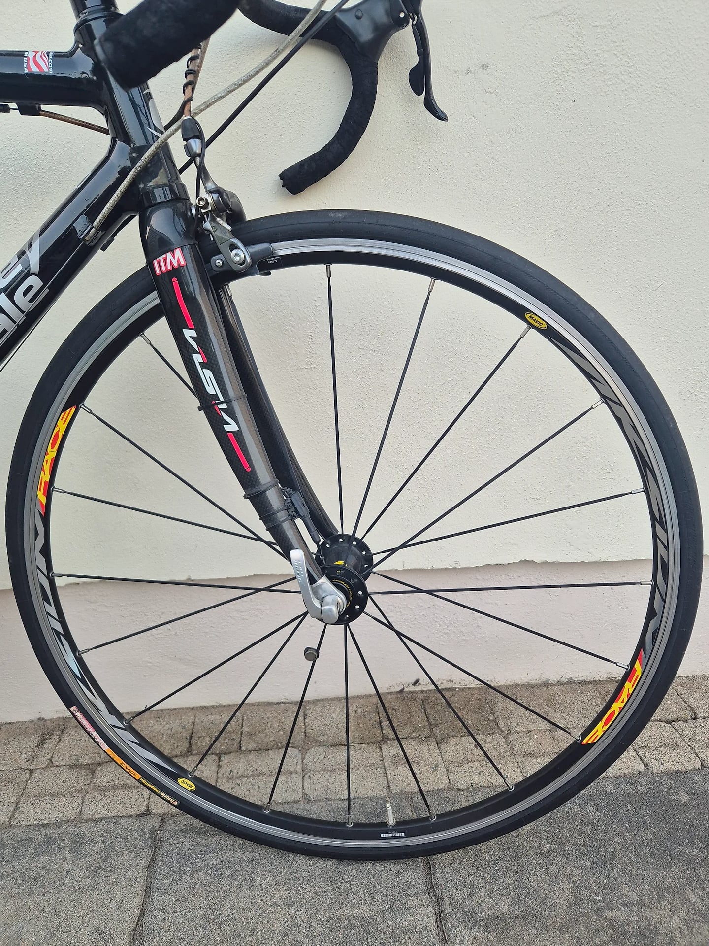 Black road bike front wheel with orange and yellow rim decals, drive side, leaning against white wall and grey pavement.