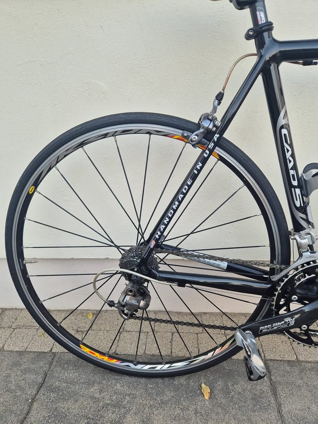 Black Cannondale road bike, front wheel close-up, drive side, against white wall and grey pavement.