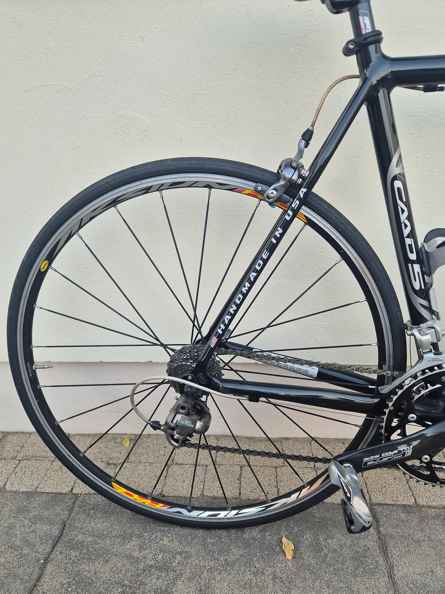 Black Cannondale road bike, front wheel close-up, drive side, against white wall and grey pavement.