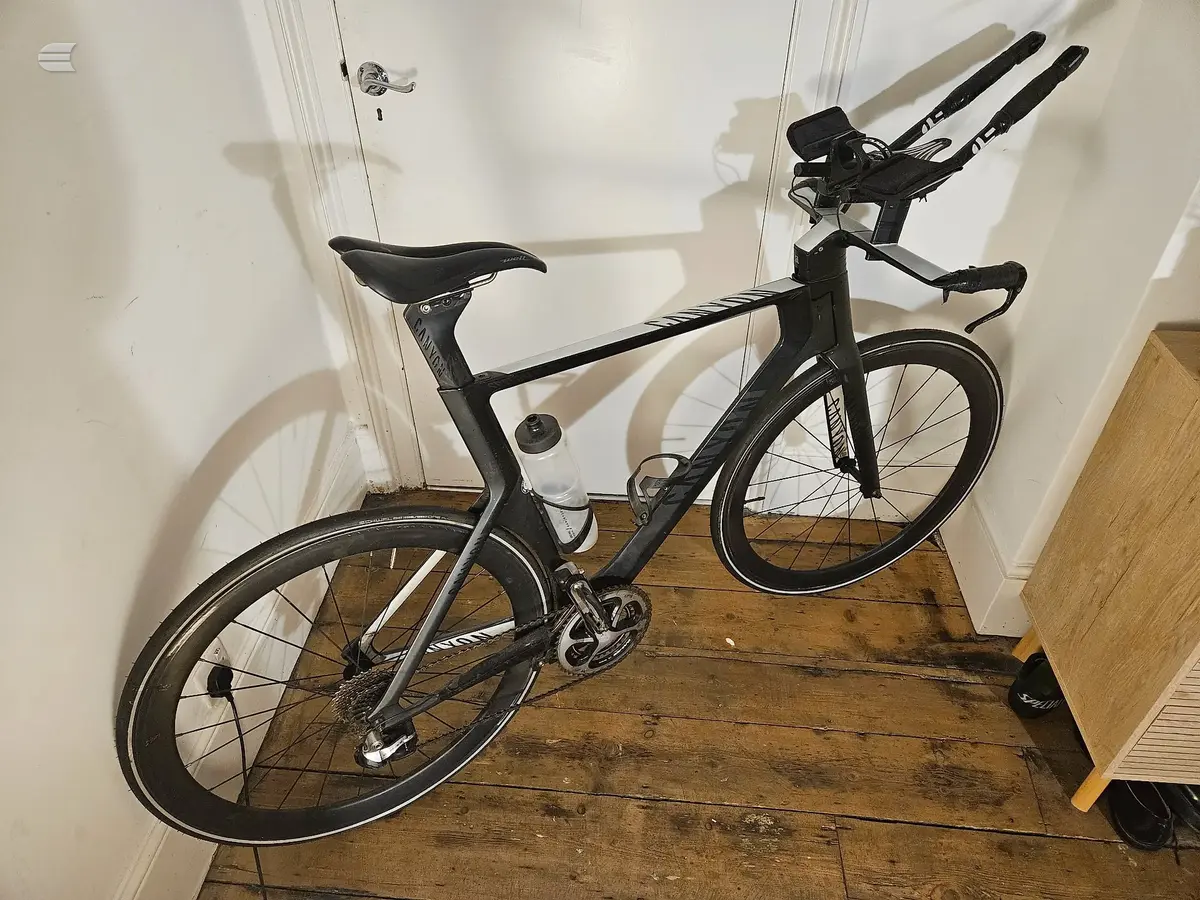 Black road bike with drop bars, indoors against white wall and wooden floor, non-drive side view, with ski poles leaning against it.