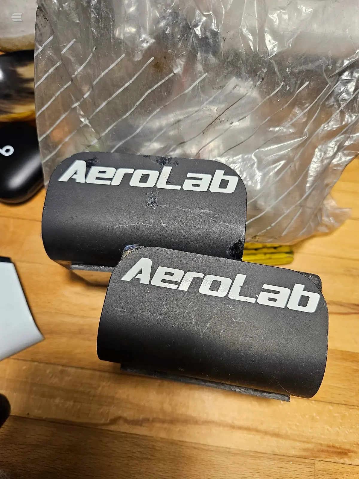 Two black AeroLab cylindrical components on wooden surface with plastic packaging behind, front view.