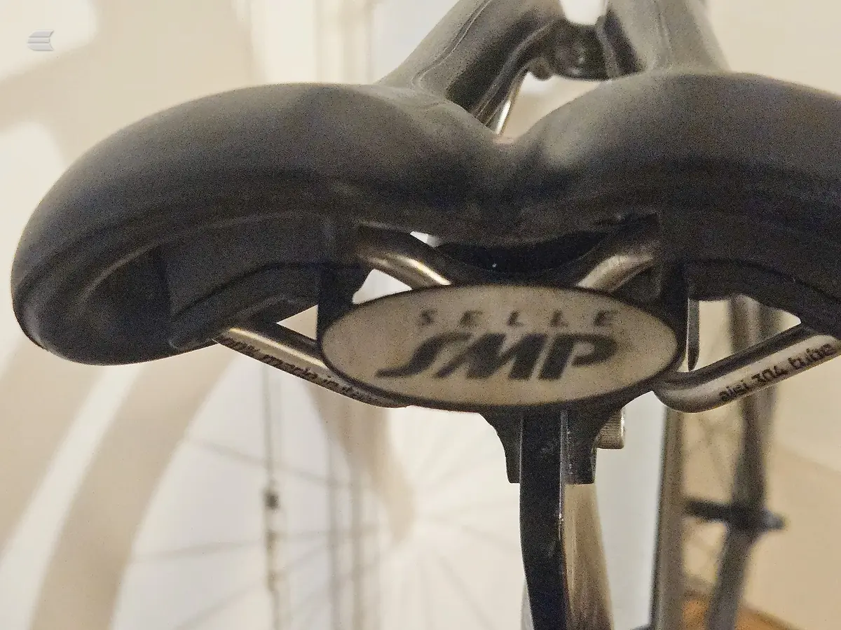 Close-up of a black bicycle saddle with 'AMP' branding visible on the seat, mounted on a metal seatpost against an indoor background.
