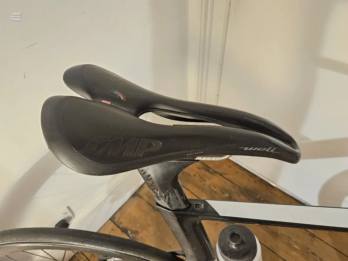 Black Selle Italia saddle mounted on bicycle seat tube, close-up view against white wall and wooden floor background.