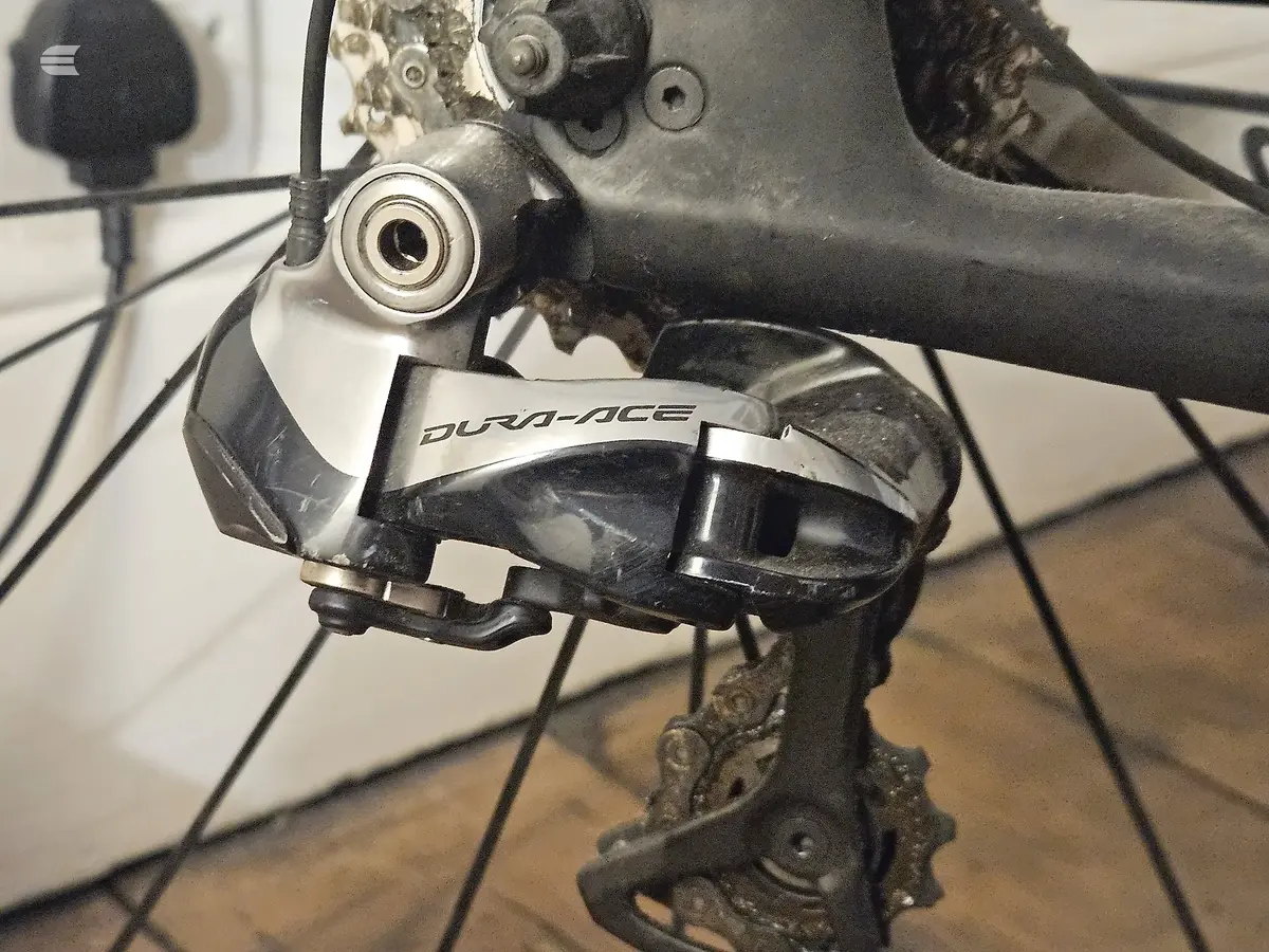 Shimano Dura-Ace rear derailleur on a road bike, drive side close-up, mounted on wooden floor indoors.