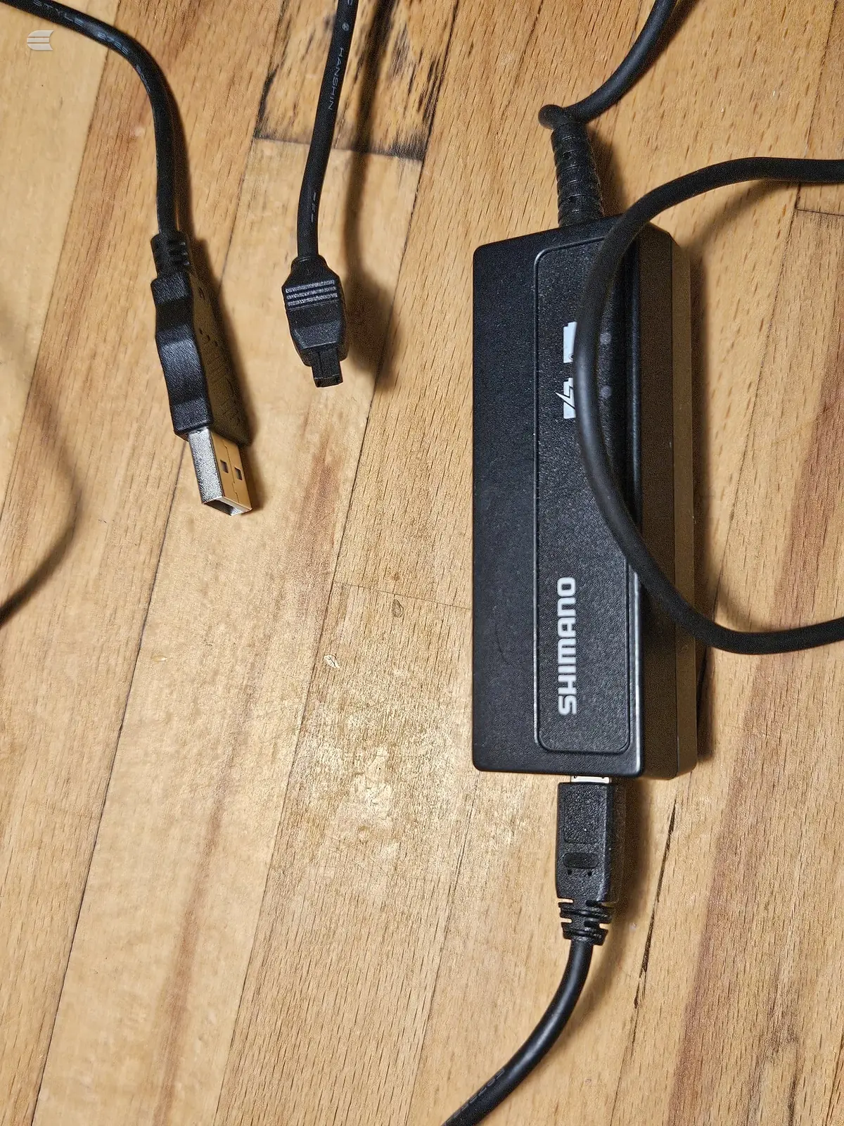 Shimano bicycle hub or component charger with USB and proprietary connectors on wooden floor.