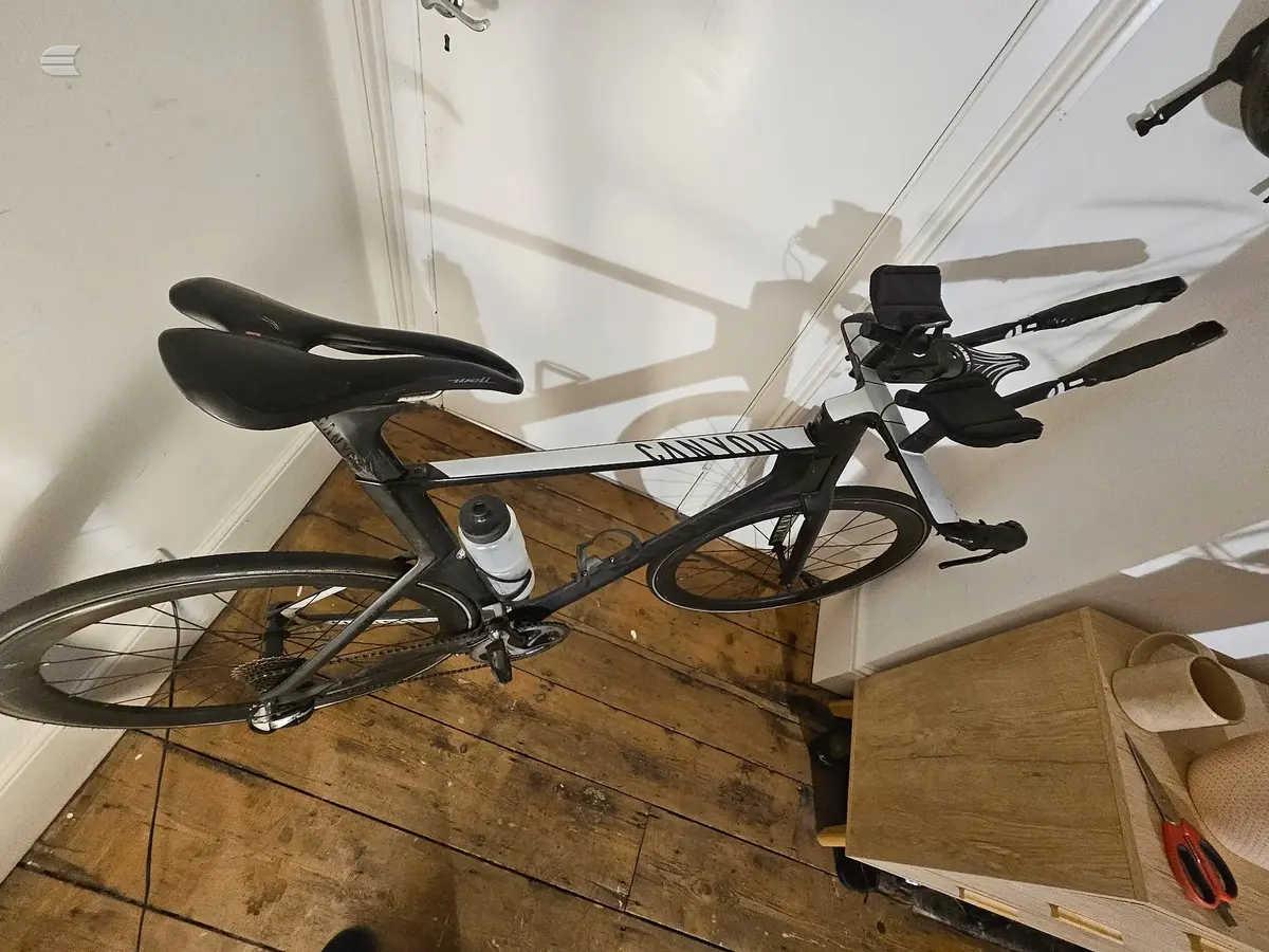 Black road bike with drop bars and aero extensions, non-drive side, on wooden floor indoors against white wall and baseboard.