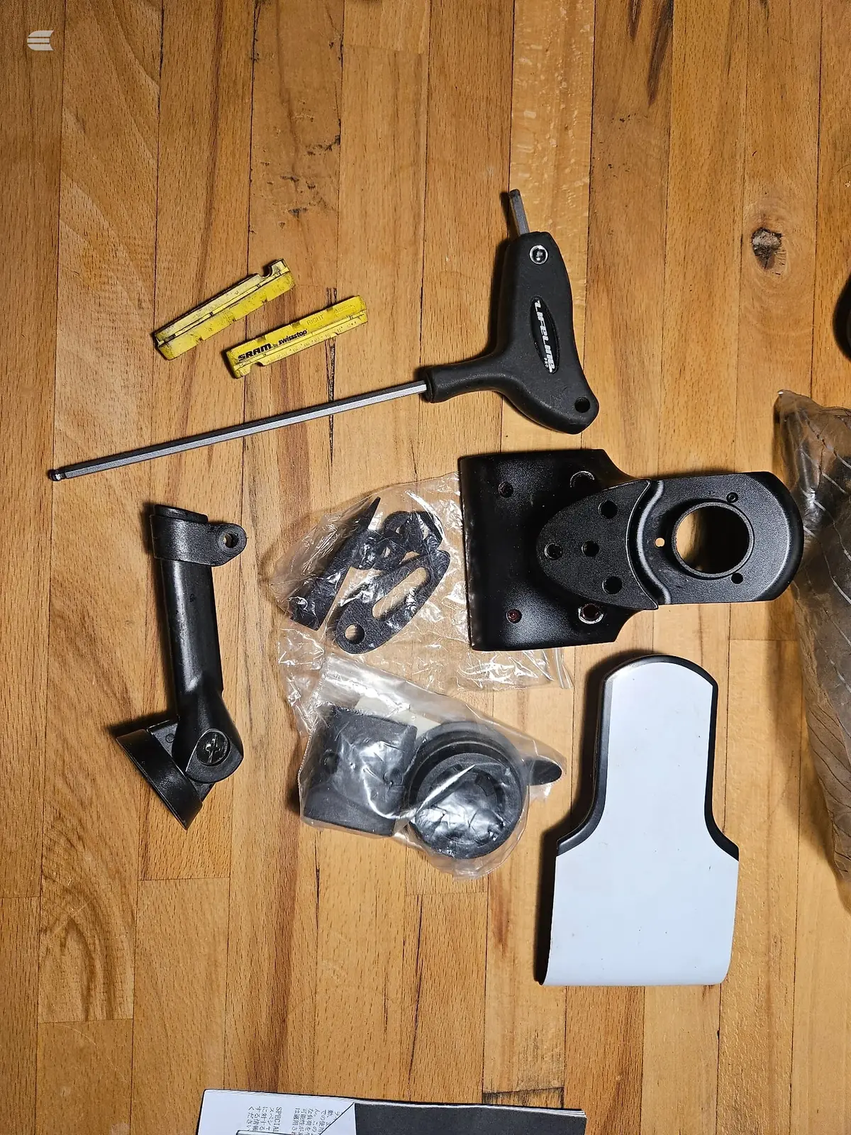Bicycle components and tools laid out on wooden floor: T-handle hex keys, spirit level, black mechanical parts in plastic bags, and white cardboard box.