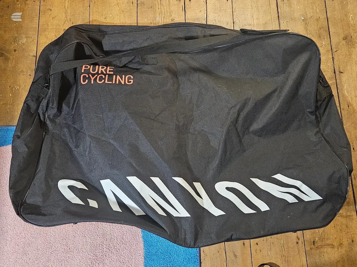 Black CANYON bike travel bag with orange 'PURE CYCLING' branding and white CANYON logo, lying flat on wooden floor with coloured fabric visible at edge.