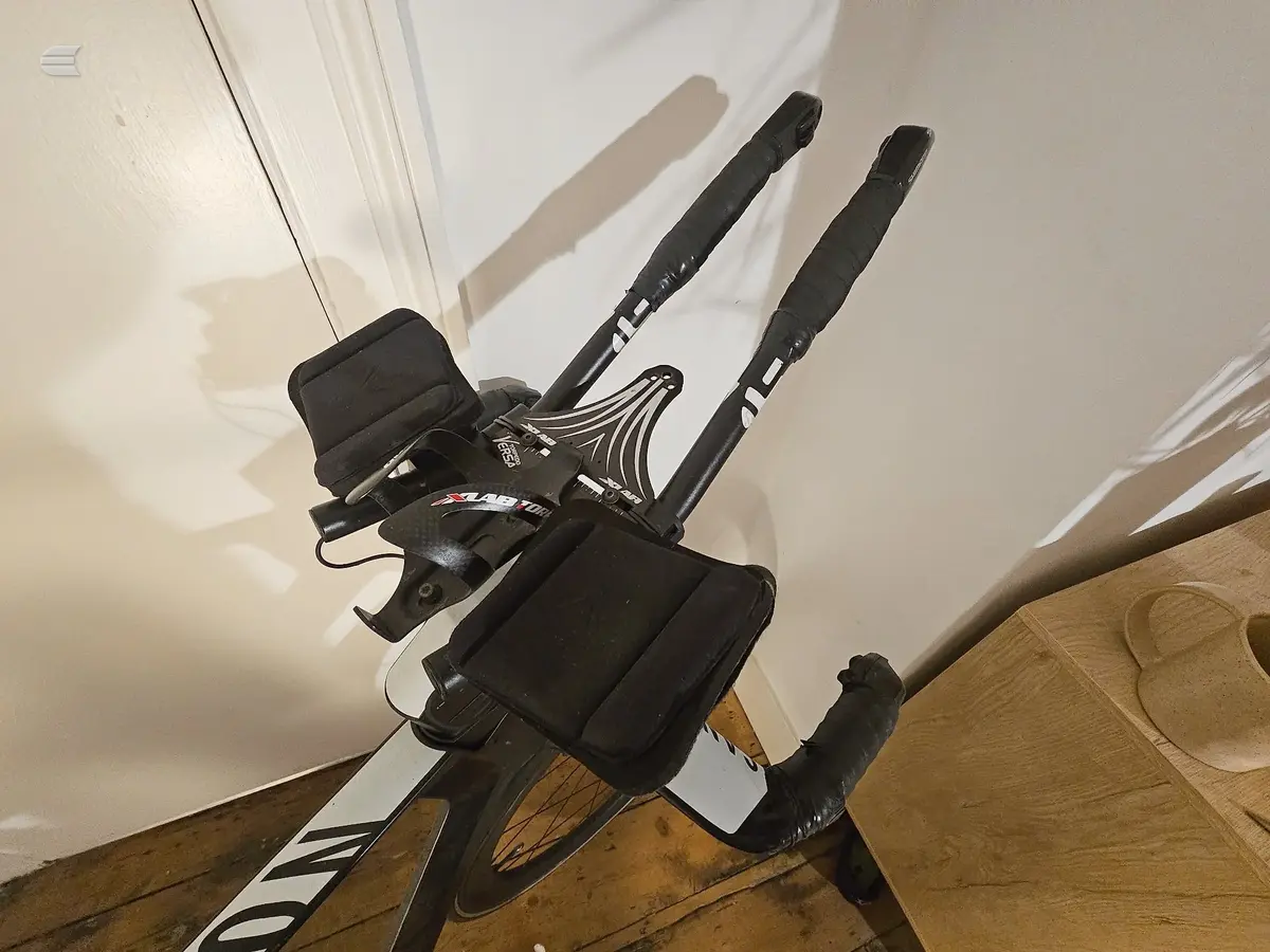 Black and white Zwift stationary bike on wooden floor indoors, non-drive side, against white wall with door frame visible.