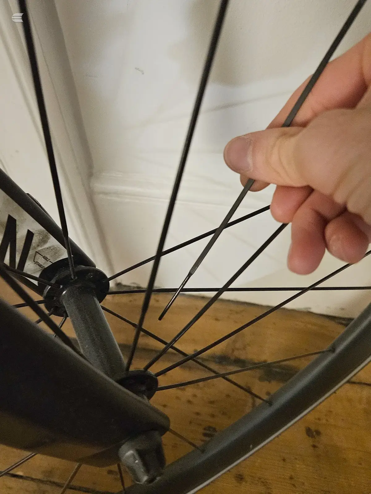 Close-up of bicycle wheel spokes and hub, hand holding spoke, wooden floor and white wall background.
